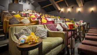 A cozy boutique cinema with tiered seating featuring plush sofas and armchairs in warm tones, decorated with colorful cushions. Small side tables hold snacks like popcorn, drinks, and candy, creating an intimate and comfortable movie-watching atmosphere.
