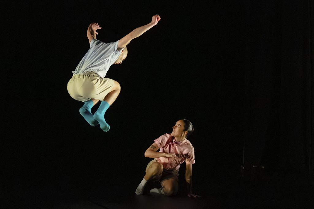 One dancer leaps into the air while another crouches below, looking up, against a dark stage background.