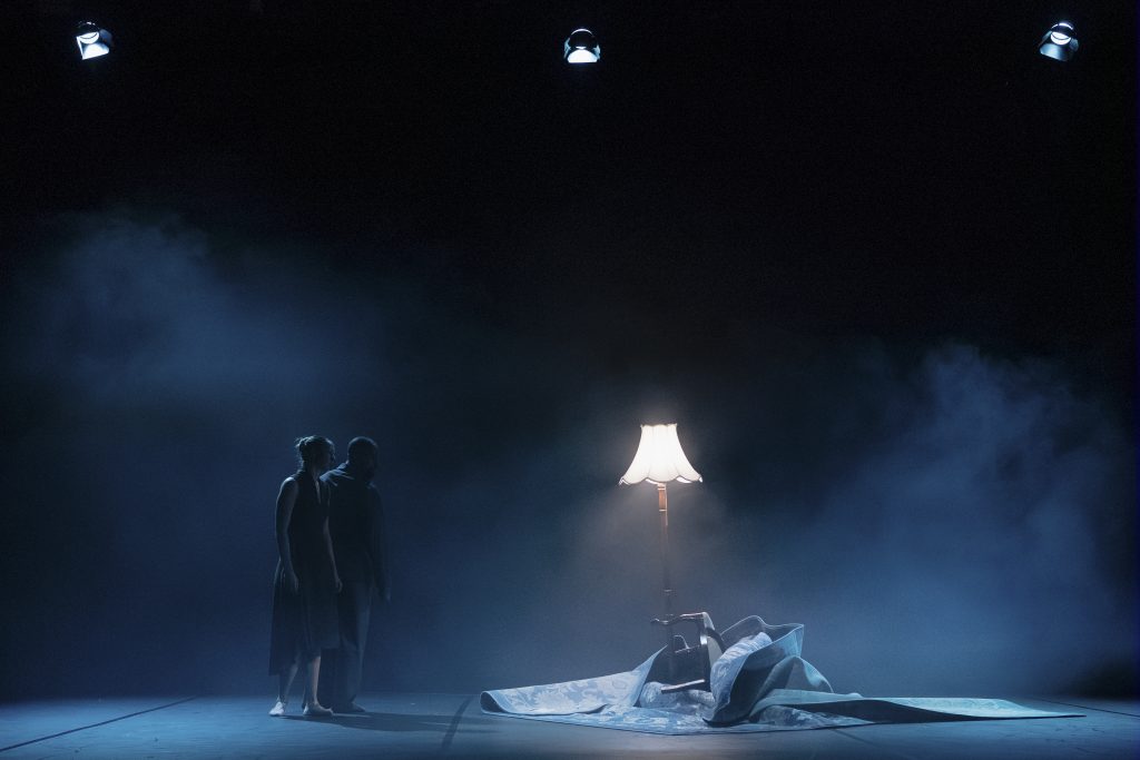 Two dancers stand side by side on a dimly lit stage, facing a softly glowing floor lamp beside an overturned chair and crumpled rug.