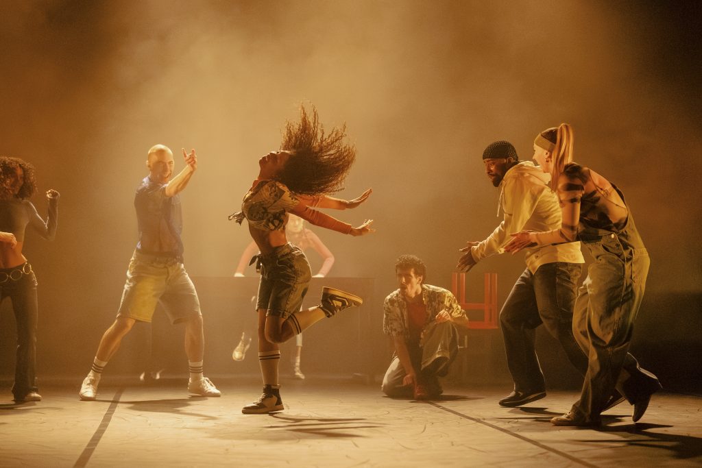 A group of contemporary dancers perform energetically on stage under warm amber lighting, with one dancer mid-jump at the centre as others gather around.