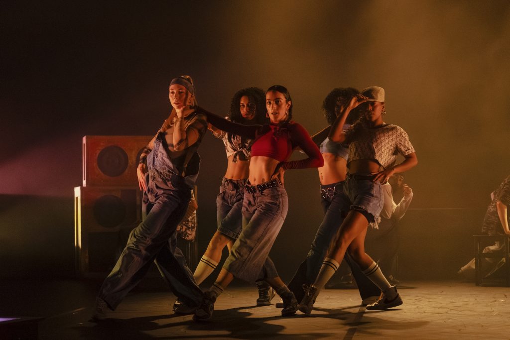 A group of dancers perform in unison under warm stage lighting, striking a stylised pose with confident, angular movements.
