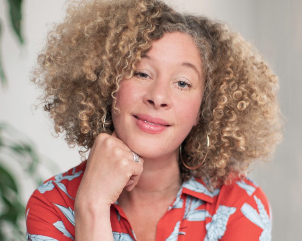 Portrait of Emily Zobel Marshall smiling gently at the camera, resting her chin on her hand. She has voluminous curly blonde hair, wears large hoop earrings and a red patterned blouse, with a softly blurred indoor background.