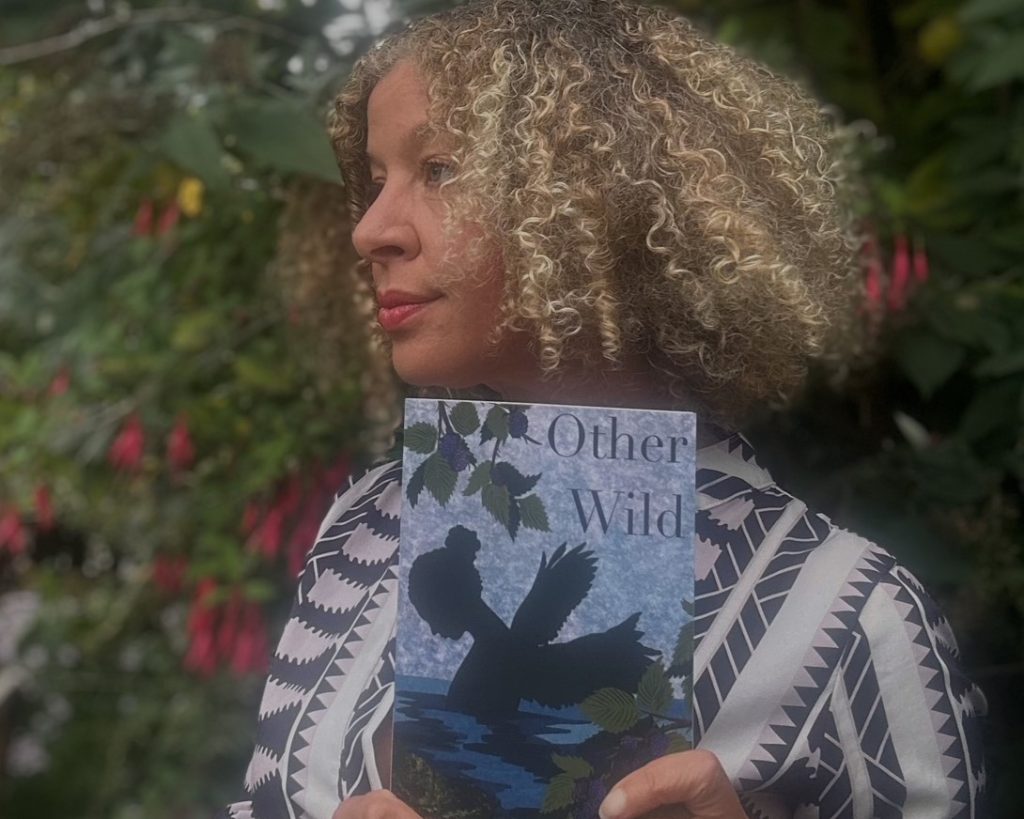A woman with curly blonde hair stands in a garden, holding a book titled “Other Wild” with a bird silhouette on the cover.