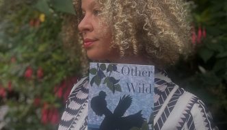 A woman with curly blonde hair stands in a garden, holding a book titled “Other Wild” with a bird silhouette on the cover.