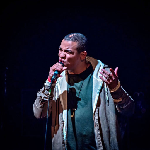 Testament performs passionately into a handheld microphone on a dark stage, wearing layered casual clothing and gesturing with one hand while singing or speaking.