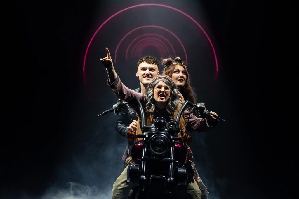 Three performers ride a motorbike on stage under circular neon lights in 'We Will Rock You.' 