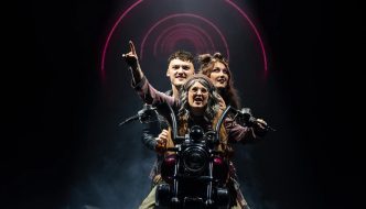 Three performers ride a motorbike on stage under circular neon lights in 'We Will Rock You.'