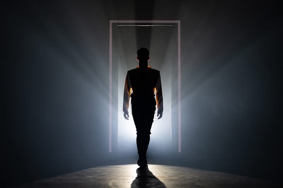 A silhouetted actor walks forward through a glowing doorway, surrounded by dramatic backlighting and haze in We Will Rock You.