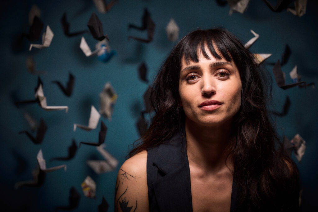 Portrait of author Rozie Kelly against a blue backdrop filled with hanging origami birds, looking directly at the camera.