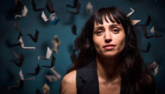 Portrait of author Rozie Kelly against a blue backdrop filled with hanging origami birds, looking directly at the camera.