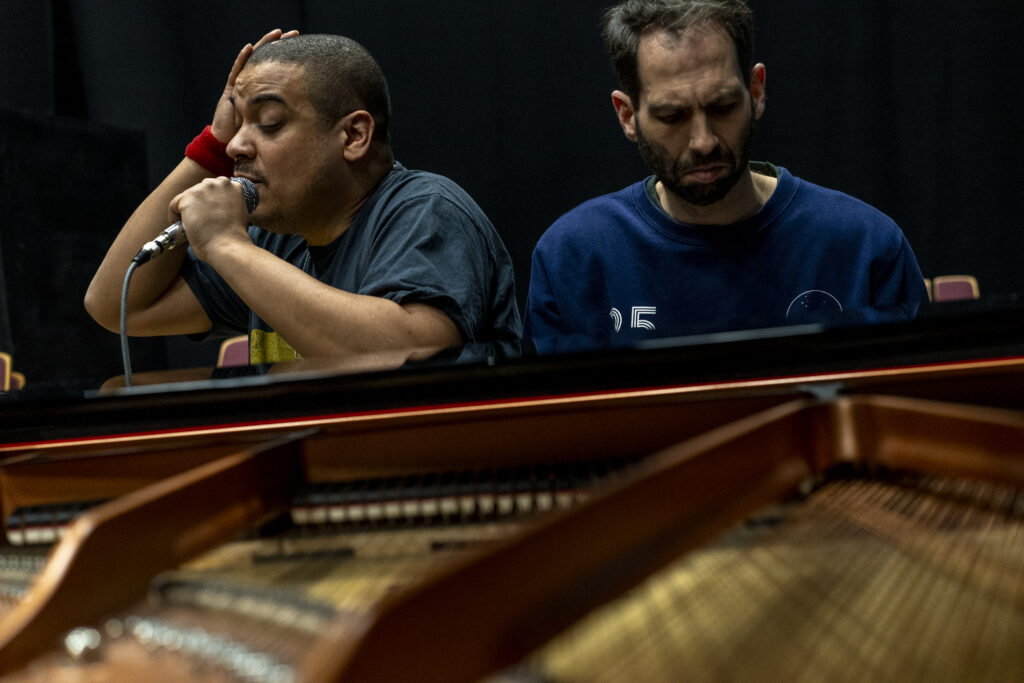 Two people sit behind a grand piano in a dark rehearsal space. One sings into a microphone while the other looks down in concentration. The piano strings fill the foreground.