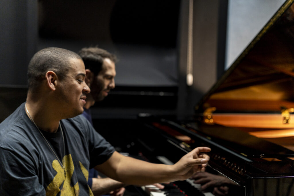Two musicians sit at a grand piano in a dimly lit room, focused on the instrument during a rehearsal or performance.