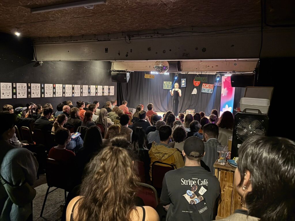 A packed audience watches a performer at Leeds Lit Fest a small, brightly lit stage in a cozy comedy venue with black curtains, colorful shop-style signs, and a disco ball overhead.