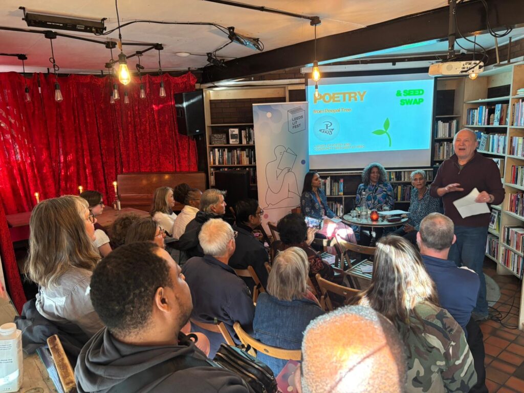 An audience looks towards a projector which says poetry and three people seated around a table.