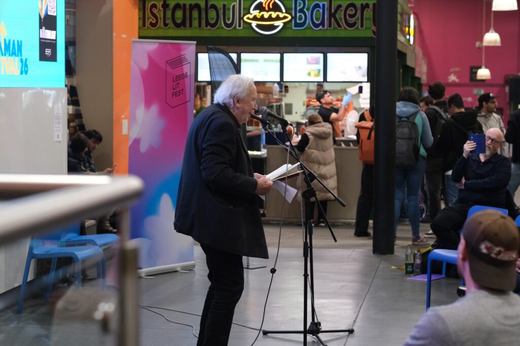 An older person reads from a book at a microphone during Leeds Lit Fest in a busy indoor market, with people watching and food stalls in the background.