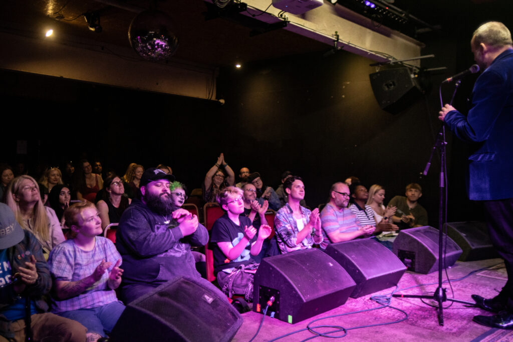 Large crowd applaud Keith Fenton stood on stage in a purple lighting.