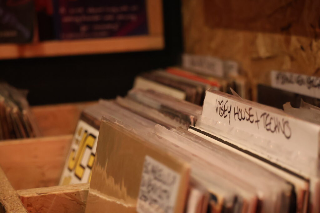 Rows of vinyl records in wooden crates at Holding Patterns, with handwritten genre dividers including "Vibey House + Techno" in sharp focus.