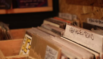 Rows of vinyl records in wooden crates at Holding Patterns, with handwritten genre dividers including "Vibey House + Techno" in sharp focus.