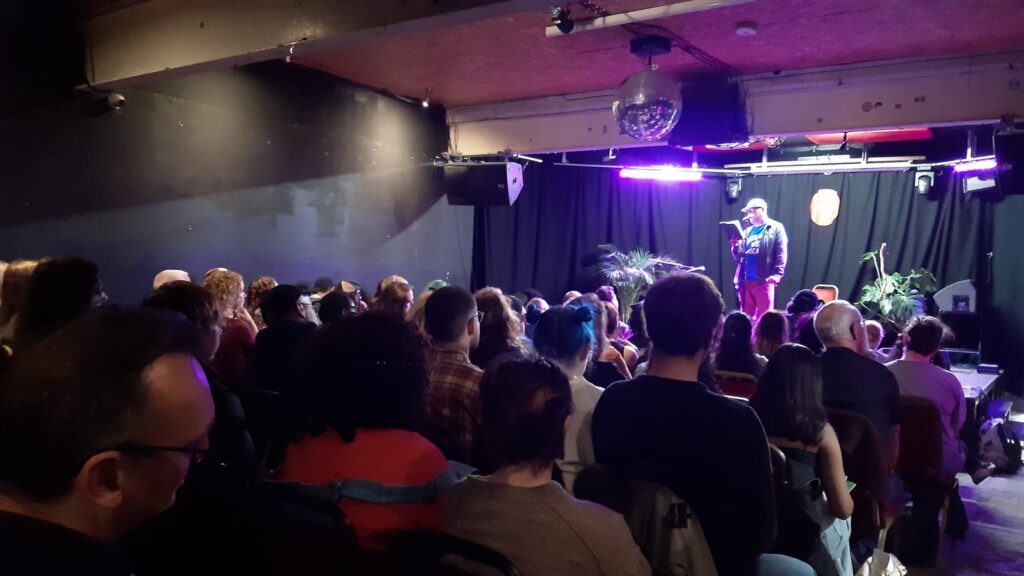 Audience watches a performer on stage in purple lighting.