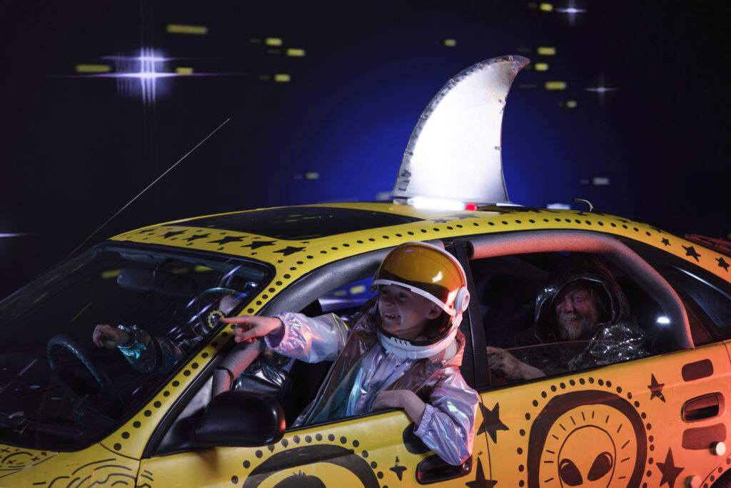 A young boy in an astronauts outfit leans out of the passenger seat of a yellow car with black paitings on pointing out of the window.