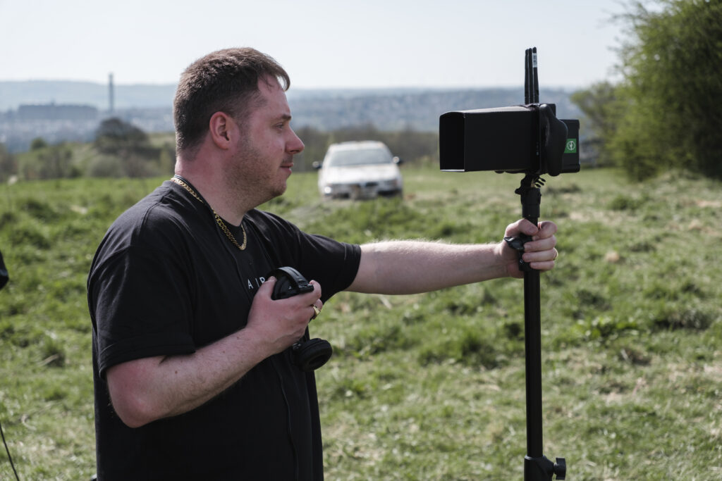 Jordon stands in a field on the set of 'Rocket Fuel' holding headphones and looking through a camera.