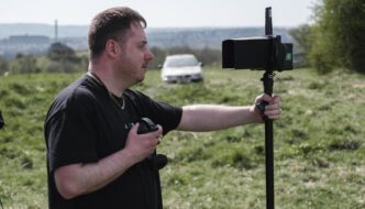Jordon stands in a field on the set of 'Rocket Fuel' holding headphones and looking through a camera.