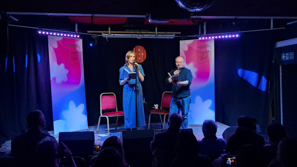 Two people read from books on stage at Leeds Lit Fest, with an audience seated in front under purple stage lights.