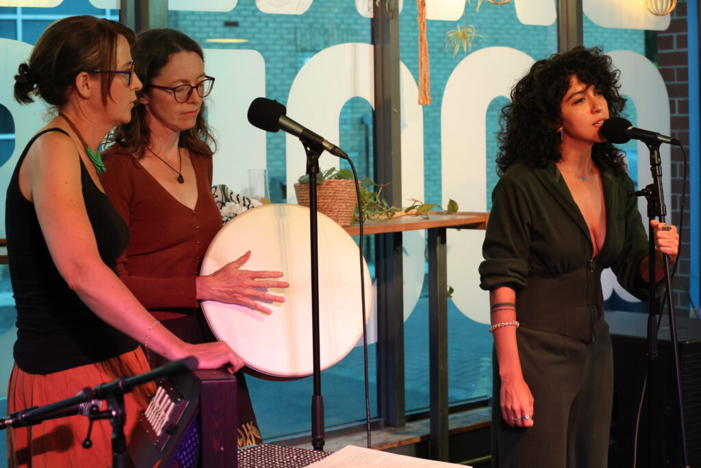 Farah Chamma performs into a microphone alongside two musicians from Adderstone, one playing a large frame drum and the other a percussion instrument.