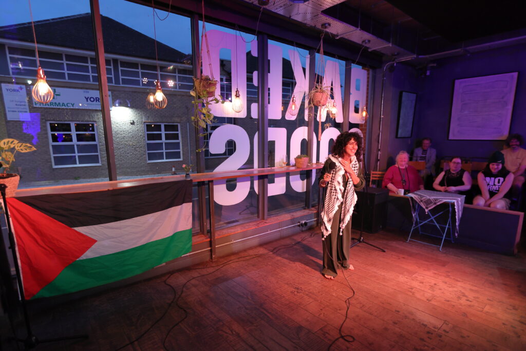Farah stood in front of a window with baked Goods written on it and a microphone in hand.