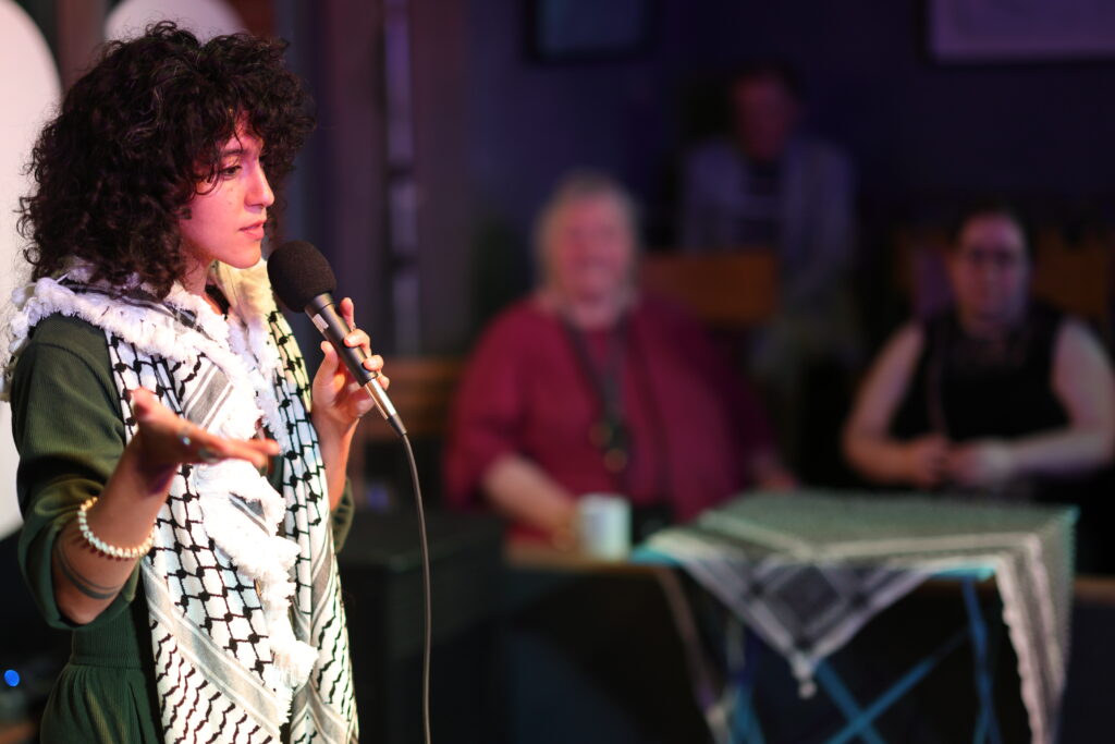 A curly-haired poet in a green outfit and black-and-white keffiyeh speaks into a microphone while gesturing with one hand; two people sit blurred in the background.