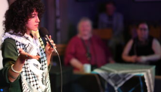 A curly-haired poet in a green outfit and black-and-white keffiyeh speaks into a microphone while gesturing with one hand; two people sit blurred in the background.