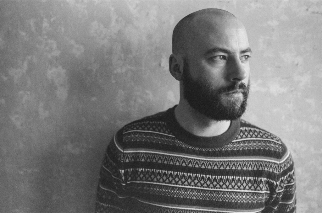 Paul Marshall, wearing a patterned jumper, is photographed in black and white against a textured wall, looking off to the side.