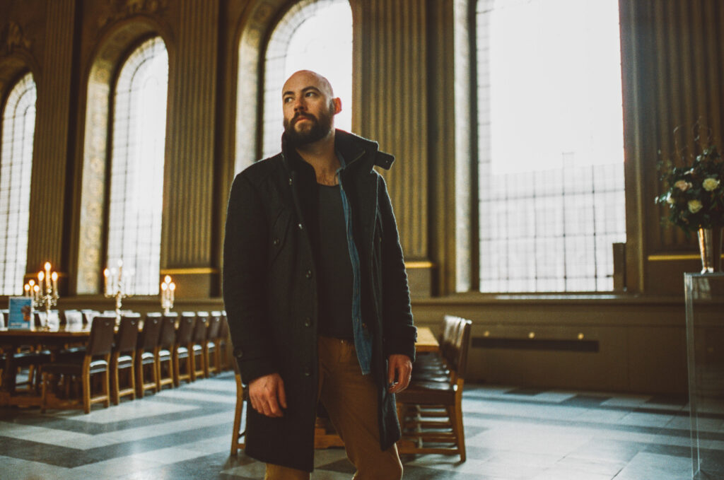 Paul Marshall, a bald, bearded man in a dark coat, stands inside a grand, warmly lit church or chapel. Tall arched windows and wooden panelling line the space, with long tables and chairs arranged behind him. He looks upward and to the side as soft daylight streams through the windows.