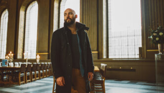 Paul Marshall, a bald, bearded man in a dark coat, stands inside a grand, warmly lit church or chapel. Tall arched windows and wooden panelling line the space, with long tables and chairs arranged behind him. He looks upward and to the side as soft daylight streams through the windows.