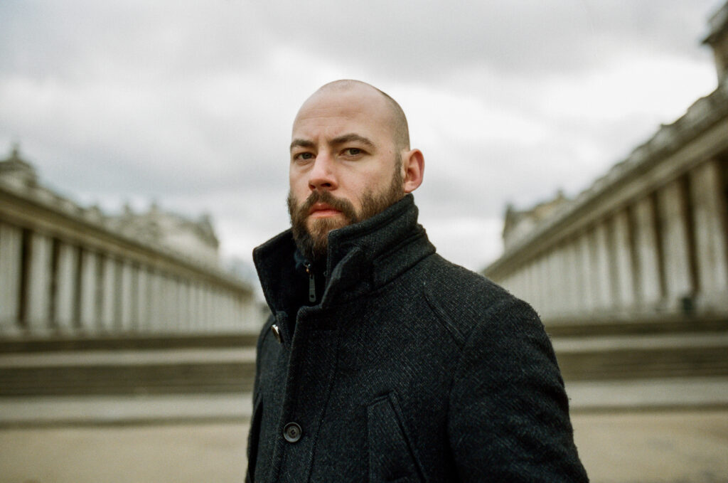 Paul Marshall stands outdoors in a dark coat, looking towards the camera. Blurred in the background are two structures supported by pillars. 