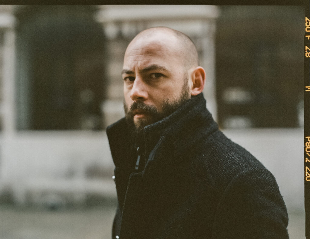 Paul Marshall stands outdoors in a dark coat and scarf, looking towards the camera. The photograph has a muted, grainy film aesthetic, with a softly blurred building in the background.