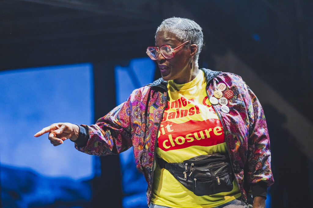 Pauline Tomlin as Rita in Brassed Off at Leeds Playhouse, wearing a bum bag, yellow t-shirt and colourful jacket with pins on. She is pointing during a performance.
