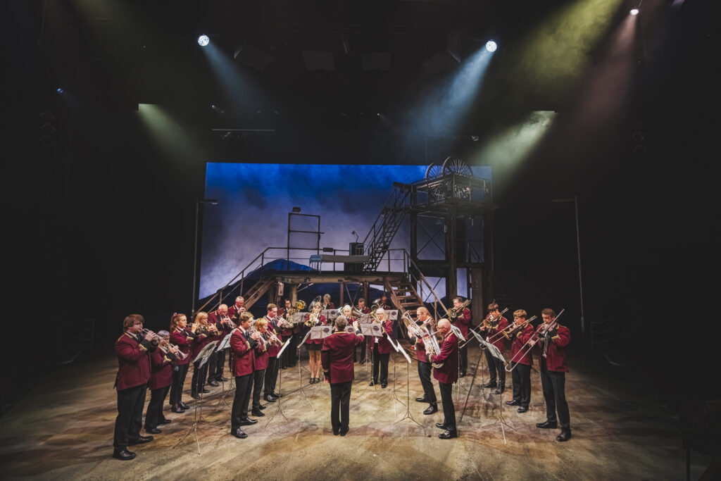 The Brassed Off company performing on stage at Leeds Playhouse, with the brass band gathered beneath the dramatic pithead-inspired set and theatrical lighting.