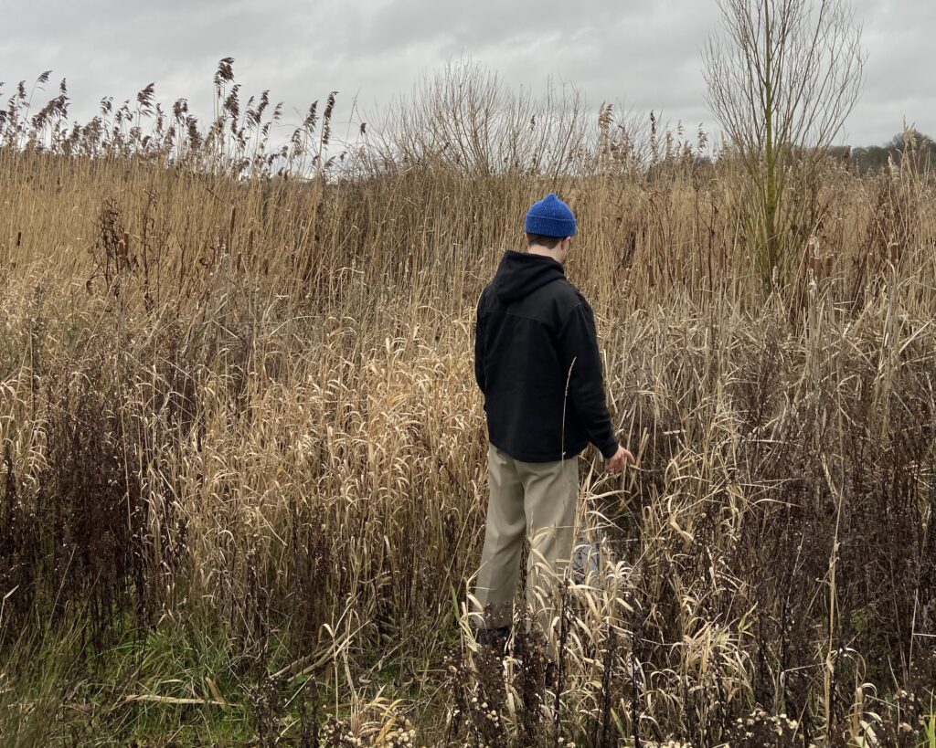 Tom stands with his back to the camera amongst some tall grass.