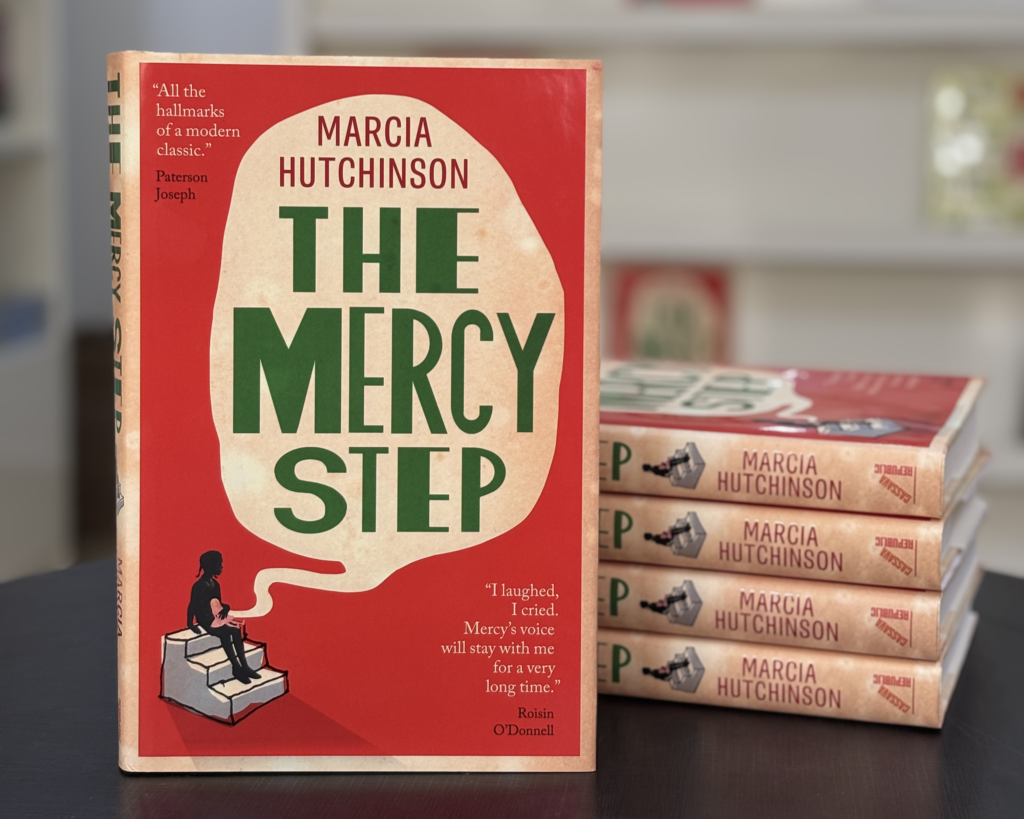 ‘The Mercy Step’ book stands on a table, leaning against four other copies. Its cover features a girl sitting on steps beneath a large thought bubble, with the title written in bold green lettering across the centre.