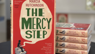 ‘The Mercy Step’ book stands on a table, leaning against four other copies. Its cover features a girl sitting on steps beneath a large thought bubble, with the title written in bold green lettering across the centre.