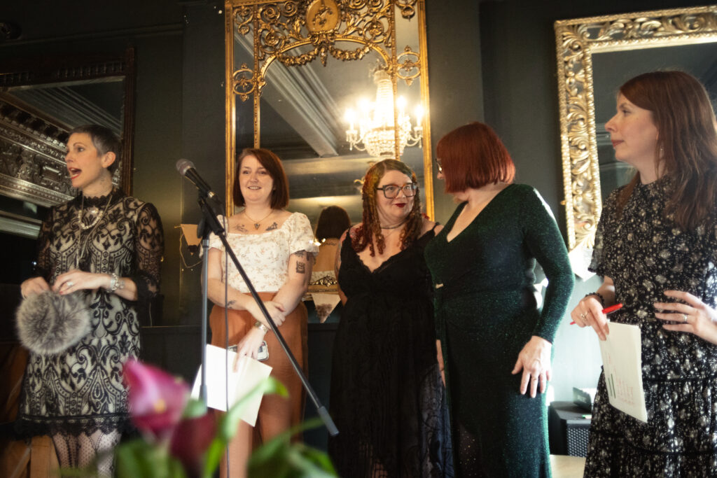 FIve women stand in a line infront of a microphone with sheets of paper in their hand.