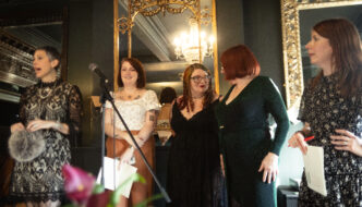 FIve women stand in a line infront of a microphone with sheets of paper in their hand.